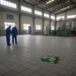Two factory workers in blue uniforms and hard hats stand in a large industrial space, observing a spilled green liquid on the acid resistant tiles Pakistan floor.